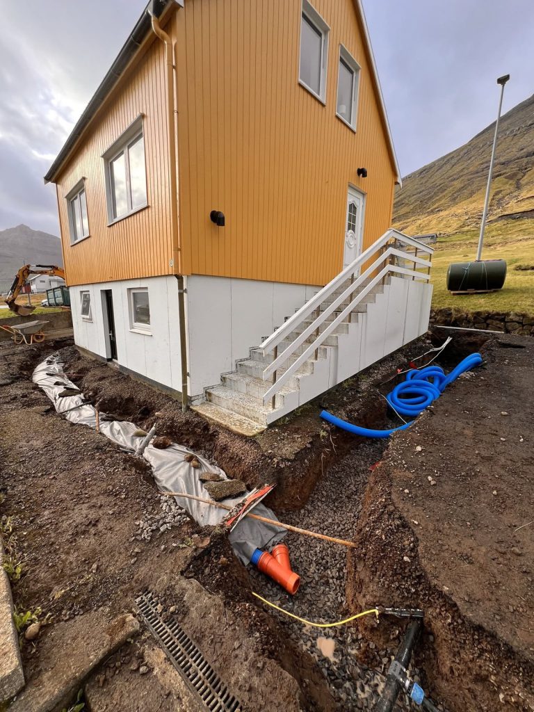 Opin dreneringsgrøv meðfram einum gulum húsi, við nýggjum rørum og dúki upp at veggin