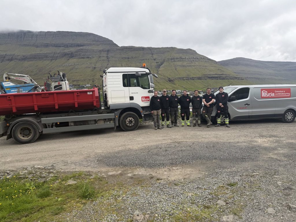 Byrja-manningin stendur millum ein reyðan tippbil og ein firmabil, við fjøllum aftanfyri