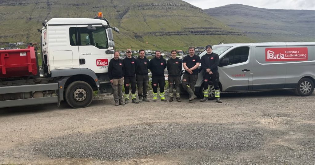 Byrja-manningin stendur á rað millum ein tippbil og ein firmabil, við fjøllum aftanfyri