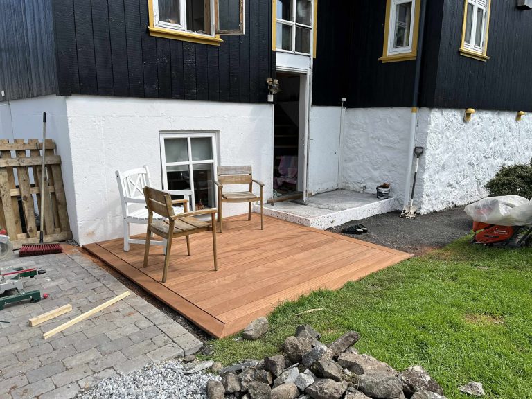 Nýggj terassa úr hørðum viði við garðstólum uttanfyri eitt svart timburhús